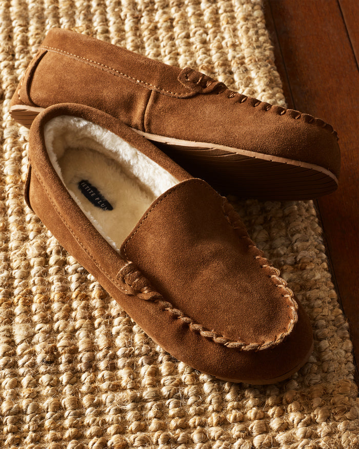 Two ultra-soft Petite Plume Womens Suede Slippers in Tan with fleece lining and detailed edge stitching rest on a textured beige rug atop a wooden floor, adding a cozy touch to the setting.