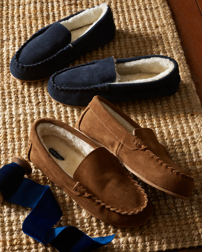 Two pairs of Petite Plumes Womens Suede Slippers in brown and navy rest on a woven mat, featuring plush faux fur lining. A spool of ribbon sits beside them.