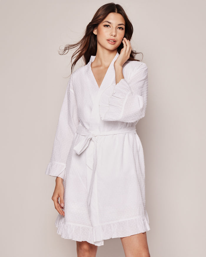 A woman models the Petite Plume Womens Swiss Dots Robe in white cotton, showcasing its ruffled cuffs and hem. She stands against a light background, touching her hair.