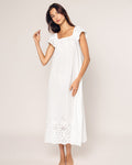 A woman stands barefoot against a plain backdrop in a sleeveless Petite Plume Womens Twill Ava Nightgown in White. Her long dark hair falls gracefully as her hands pose, highlighting the nightgowns intricate patterns at the hem and neckline.
