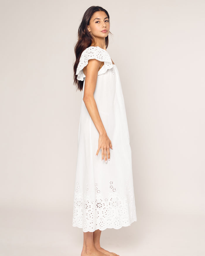 A woman in a white, off-shoulder dress gazes left while standing barefoot on a neutral background. Shes wearing the Womens Twill Ava Nightgown by Petite Plume, featuring an ankle-length silhouette crafted from fine cotton with eyelet details.