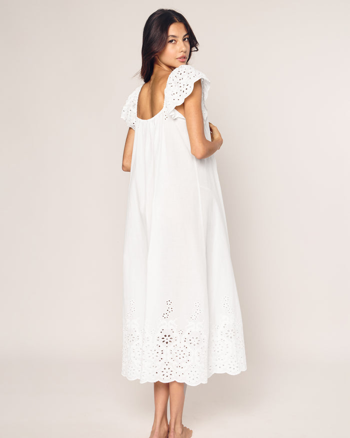 A person stands barefoot, facing away, in a white sleeveless Womens Twill Ava Nightgown by Petite Plume, adorned with intricate eyelet embroidery on the hem and shoulder frills. Made from premium cotton, this elegant sleepwear beautifully contrasts with the plain off-white background.