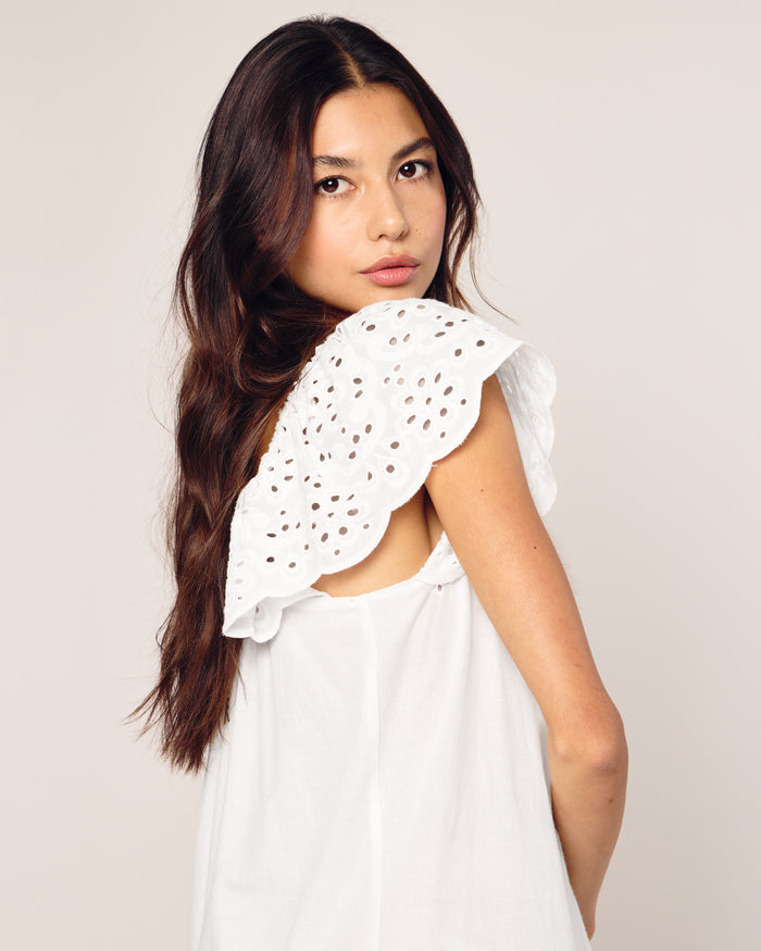 A woman with long, dark hair wears a Womens Twill Ava Nightgown in White from Petite Plume. The dress features delicate lace and ruffled shoulder details. She looks over her shoulder with a neutral expression against a plain, light-colored background.