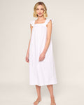 A blonde woman stands barefoot in a Womens Twill Clara Nightgown by Petite Plume, a calf-length white dress with ruffled shoulders. Made from premium cotton, the luxurious sleepwear is set against a plain light-colored background.