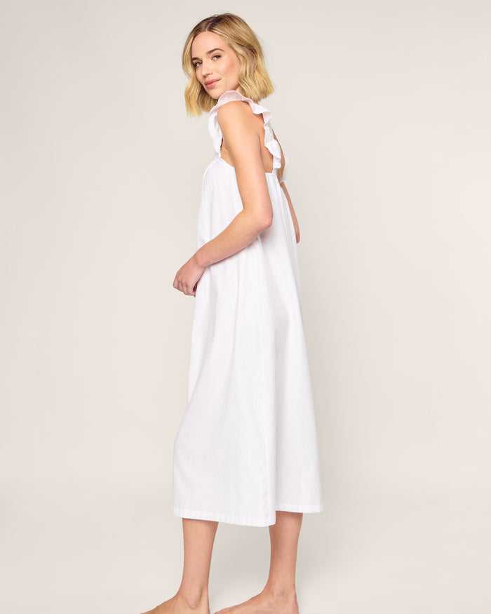 A woman in a white, sleeveless Womens Twill Clara Nightgown by Petite Plume stands barefoot on a light background. She is turned sideways, smiling at the camera. The nightgown features ruffled straps and an open back, crafted from high-quality cotton for luxurious sleepwear.