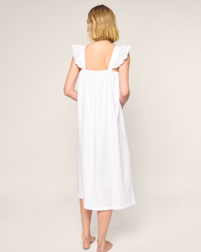 A person with short blonde hair stands barefoot on a neutral background, wearing the luxurious Womens Twill Clara Nightgown in White by Petite Plume. This sleeveless, knee-length dress has lace detailing on the shoulders and back, crafted from top-quality cotton. The back is visible.