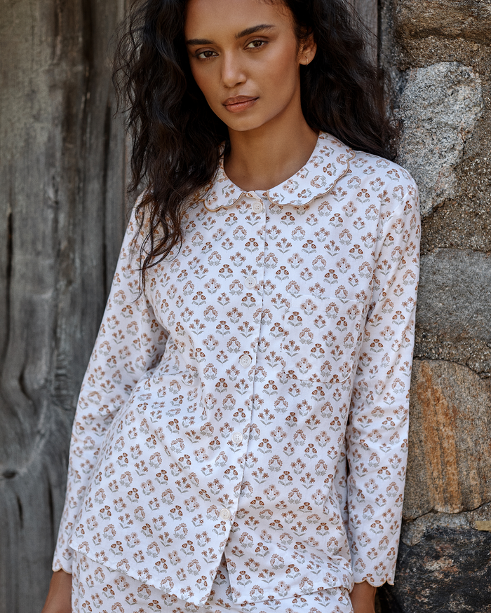 A woman with long dark hair wears the Women’s Twill Colette Pajama Set in Estate Garden, standing in front of a rustic wooden and stone wall and looking at the camera with a relaxed expression.