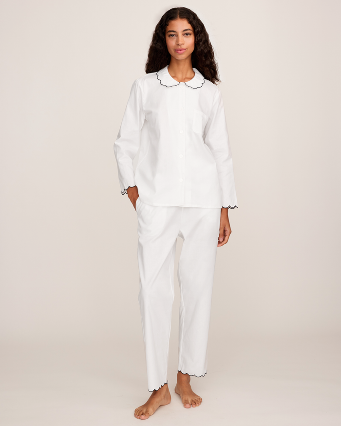 A person stands barefoot against a plain light background, wearing the Women’s Twill Colette Pajama Set in White with Navy Piping, which features contrasting trim on the collar, sleeves, and pant cuffs.