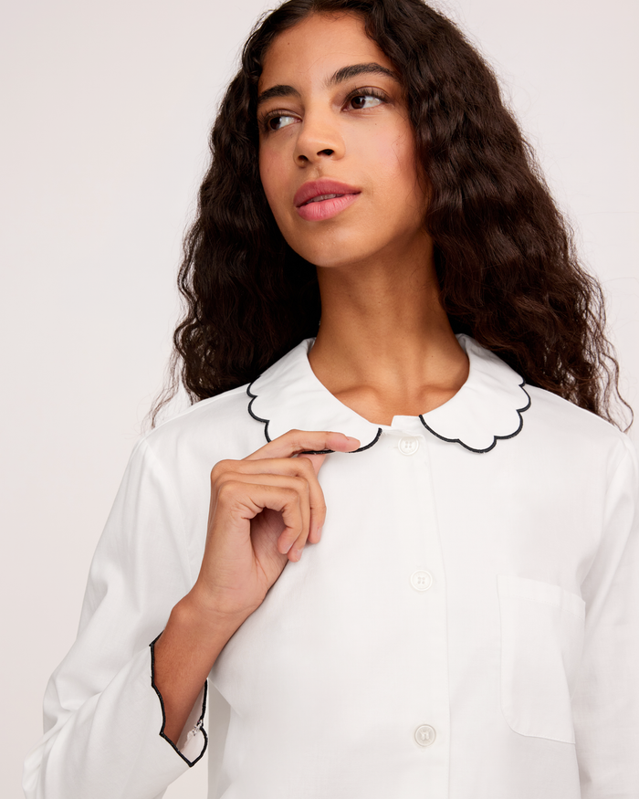 A woman with long, wavy hair wears the Women’s Twill Colette Pajama Set in White with Navy Piping, looking slightly to the side while holding the edge of her collar.