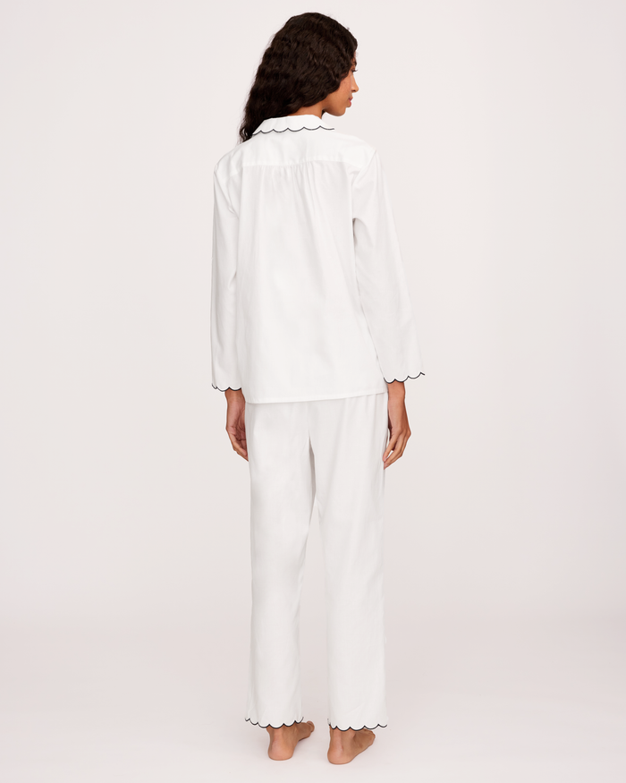 A person stands barefoot facing away, wearing the Women’s Twill Colette Pajama Set in White with Navy Piping, which features long sleeves and navy-trimmed edges on the collar, sleeves, and pant legs. The background is plain and light-colored.