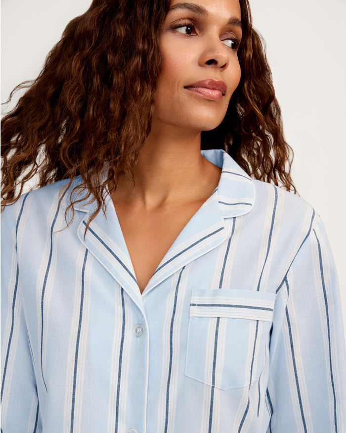 A woman with wavy brown hair wears the Women’s Twill Nightshirt in Coastal Stripe—a light blue and white striped button-up shirt with a collar and chest pocket—while glancing slightly to the side against a plain background.