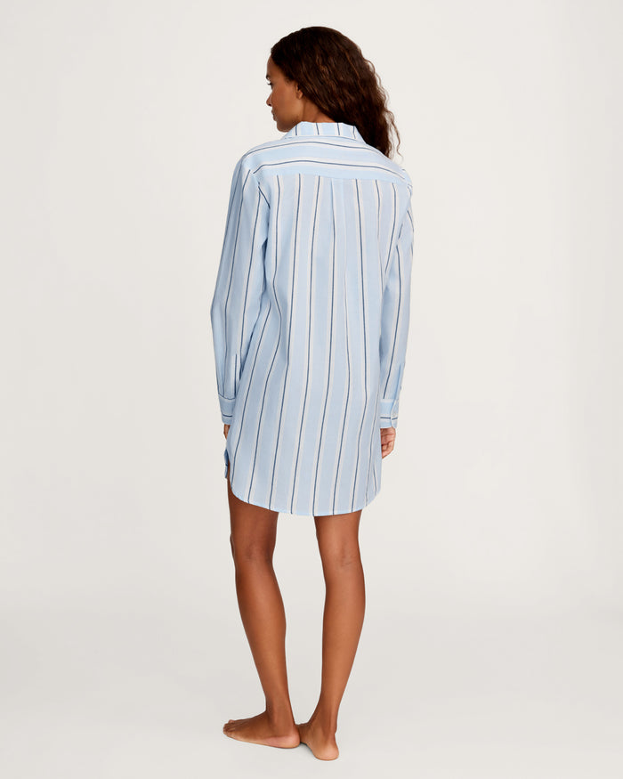 A woman stands barefoot, facing away, wearing the Women’s Twill Nightshirt in Coastal Stripe—a long-sleeved, light blue nightshirt featuring vertical white and dark stripes. The background is plain and light-colored.