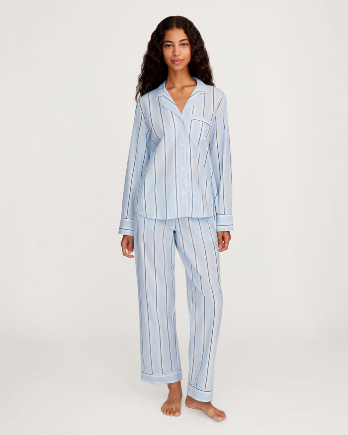 A woman stands barefoot against a plain white background wearing the Women’s Twill Pajama Set in Coastal Stripe—light blue, long-sleeved, button-up pajamas featuring vertical white and dark stripes.