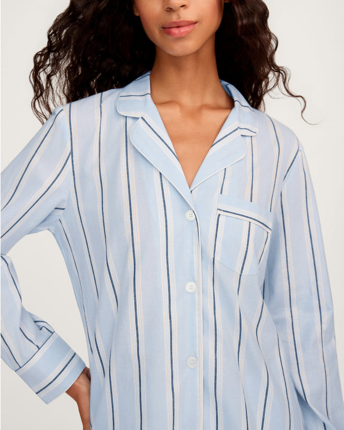 A woman stands facing forward with one hand on her hip, wearing the Women’s Twill Pajama Set in Coastal Stripe—light blue, white, and navy striped pajamas featuring a collar and chest pocket.
