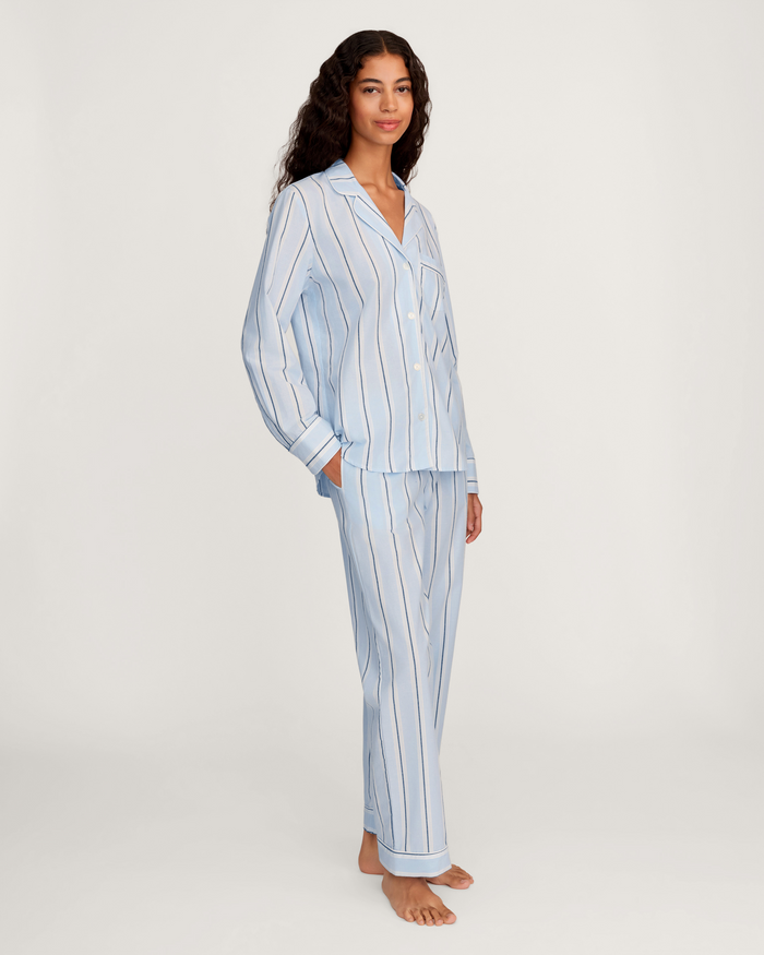 A woman stands barefoot on a plain background, wearing the Women’s Twill Pajama Set in Coastal Stripe—a light blue, long-sleeved, button-up style featuring vertical black and white stripes.