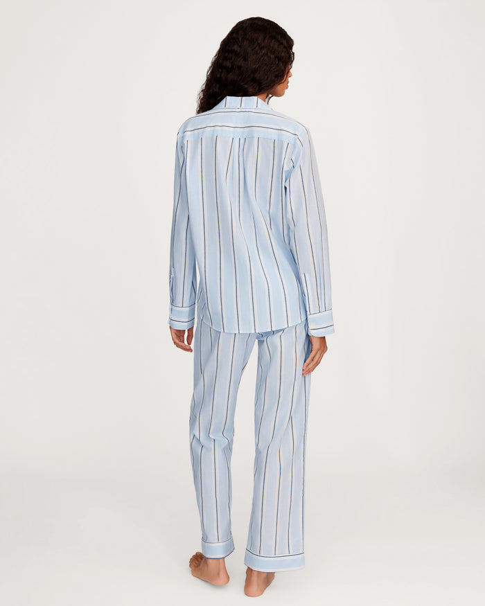 A person stands barefoot facing away, wearing the Women’s Twill Pajama Set in Coastal Stripe—a light blue pajama set with vertical white and dark stripes, featuring a long-sleeve top and matching pants, against a plain white background.