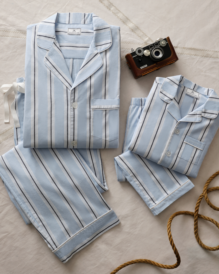Two Women’s Twill Pajama Sets in Coastal Stripe are neatly folded on a white cloth. Nearby, a vintage camera and a coiled brown rope add a nostalgic touch to the scene.