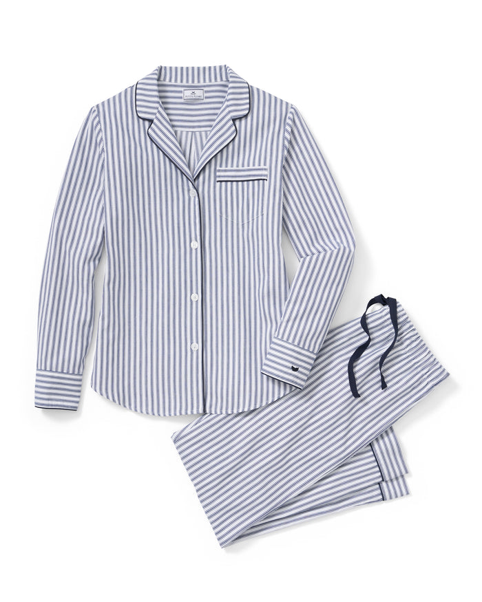 A model with long hair, wearing the Petite Plume Womens Twill Pajama Set in Navy French Ticking, poses against a plain background with hands in pockets. The cotton top features a collar and the pants are full-length, complementing her relaxed expression.