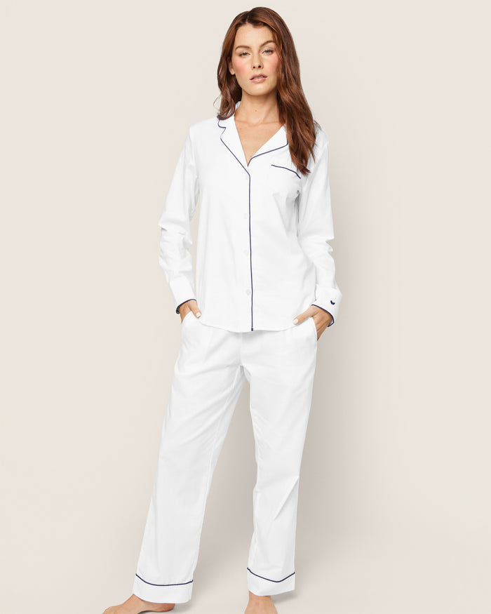 A person in Petite Plumes Womens Twill Pajama Set in white with navy piping stands against a plain background. The top has a collar and buttons, and the loose-fitting, moisture-wicking pants have pockets. They hold a neutral expression with one hand in their pocket.