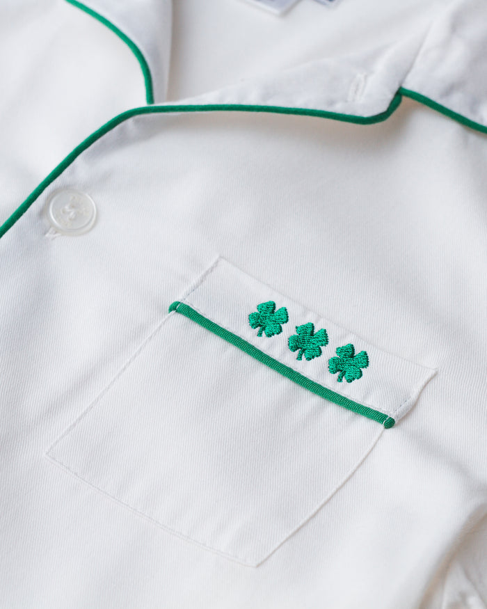 Close-up of a white fabric from the Petite Plume Womens Twill Pajama Set, featuring a pocket with green trim and three embroidered shamrock symbols for a St. Patricks Day touch. The fabric is smooth, with a button partially visible.