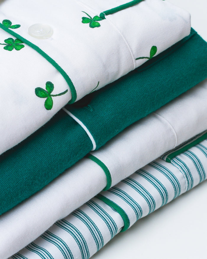 A neatly folded stack of the Petite Plume Womens Twill Pajama Set features: a white top with green shamrock embroidery, a solid green shirt, a plain white layer, and one with green stripes. Perfect for a festive St. Patricks Day look!.