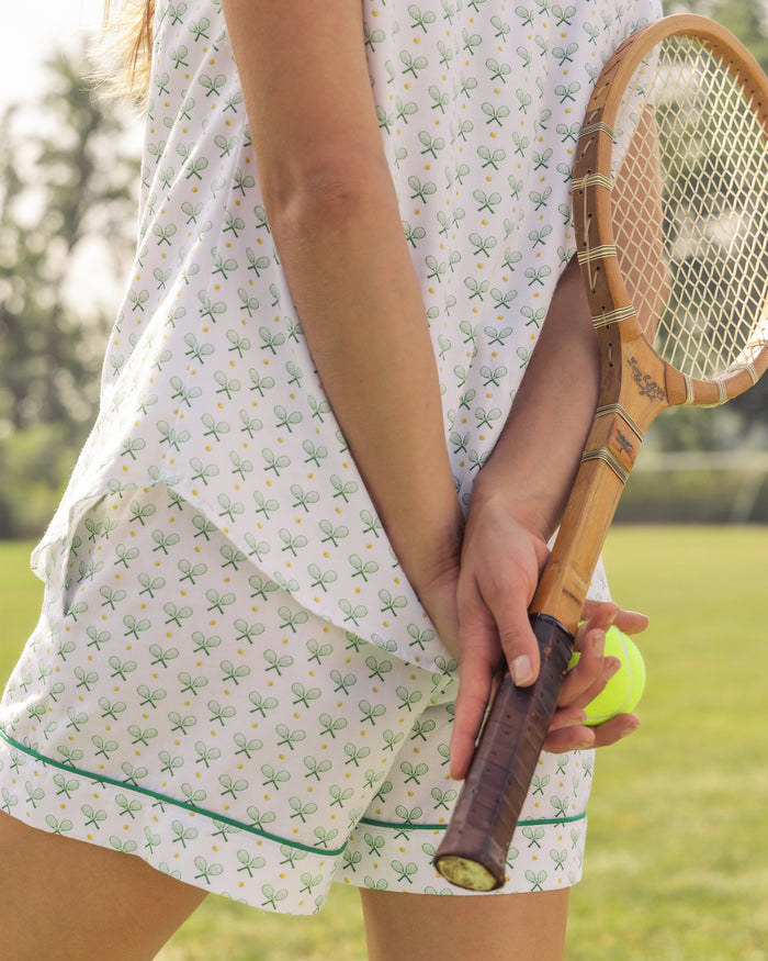 A person in a white outfit with a green tennis racket print, reminiscent of luxury pajamas, stands on a grassy field holding a wooden tennis racquet and ball. Trees enhance the serene scene. Shes wearing the Womens Twill Pajama Short Sleeve Short Set in Match Point by Petite Plume.
