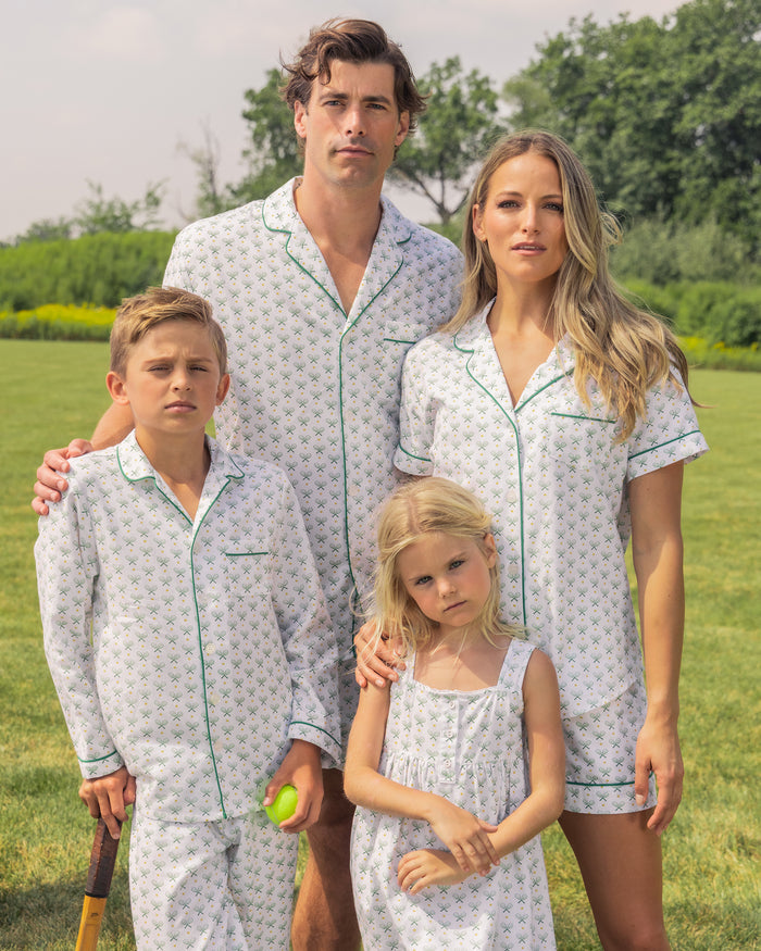 A family of four poses outdoors in luxury pajamas. The mom wears a Womens Twill Pajama Short Sleeve Short Set in Match Point by Petite Plume. An adult male, young boy with a bat and ball, and young girl in a dress join her on grassy terrain amidst trees, relishing their stylish sleepwear moment.