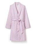 A woman stands barefoot against a plain light background, wearing the Women's Twill Robe in Fleur Blush—a knee-length, long-sleeved, light pink robe with a subtle floral pattern and waist tie.
