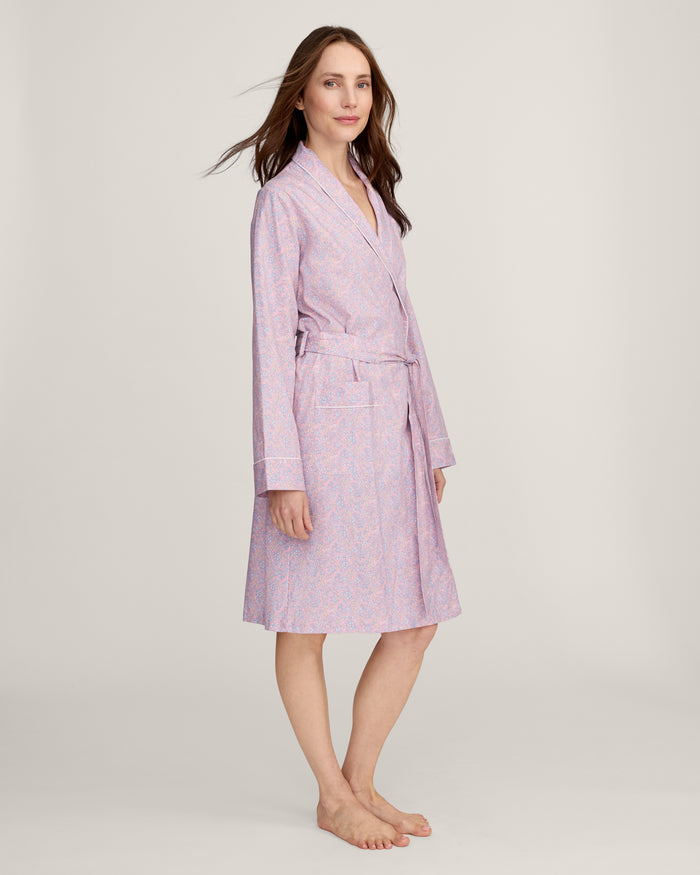 A woman stands barefoot against a plain light background, wearing the Women's Twill Robe in Fleur Blush—a knee-length, long-sleeved, light pink robe with a subtle floral pattern and waist tie.