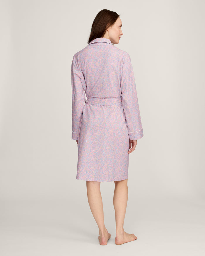 A woman stands barefoot against a plain light background, wearing the Women's Twill Robe in Fleur Blush—a knee-length, long-sleeved, light pink robe with a subtle floral pattern and waist tie.