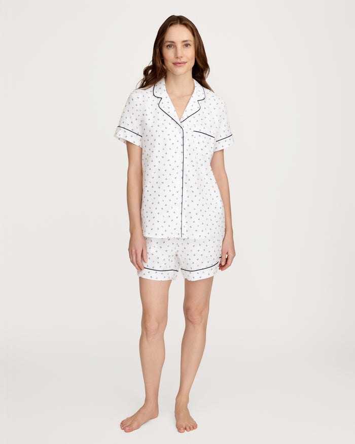 A woman stands barefoot against a plain light background, wearing the Women’s Twill Short Sleeve Short Set in Anchors Away White—a white pajama set with short sleeves and shorts, featuring a small anchor pattern and dark piping.