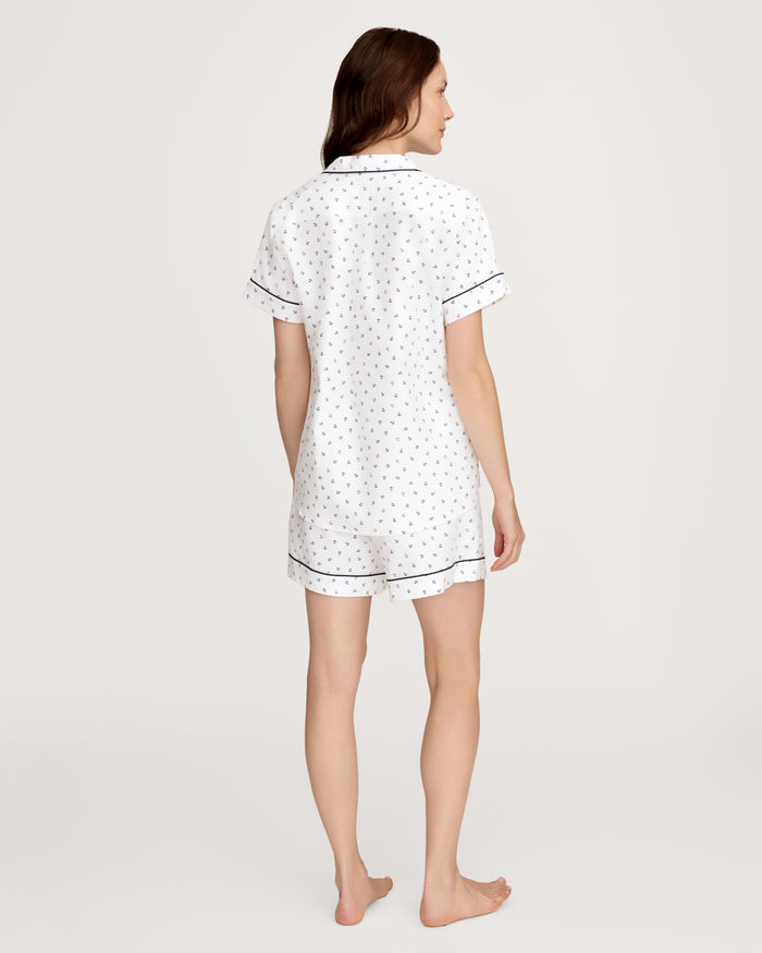 A woman stands barefoot facing away, wearing the Women’s Twill Short Sleeve Short Set in Anchors Away White, which features a subtle dotted pattern and black piping, against a plain light background.