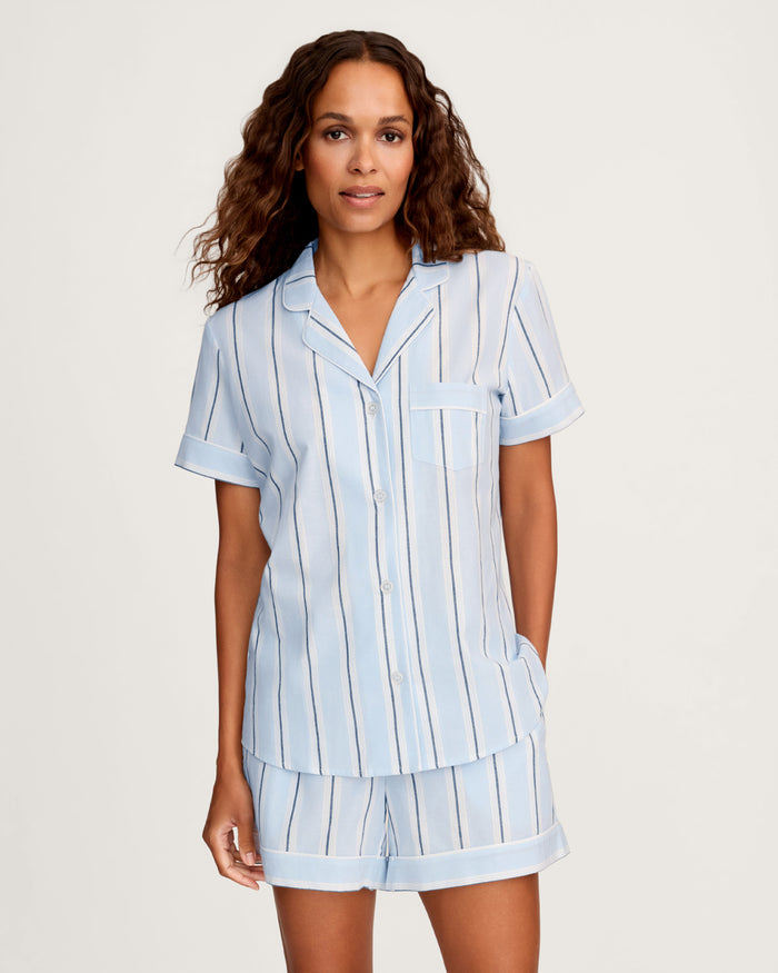 A woman wears the Women’s Twill Short Sleeve Short Set in Coastal Stripe, featuring a light blue, short-sleeved, button-up pajama top and matching shorts with dark blue and white vertical stripes, standing against a plain background.