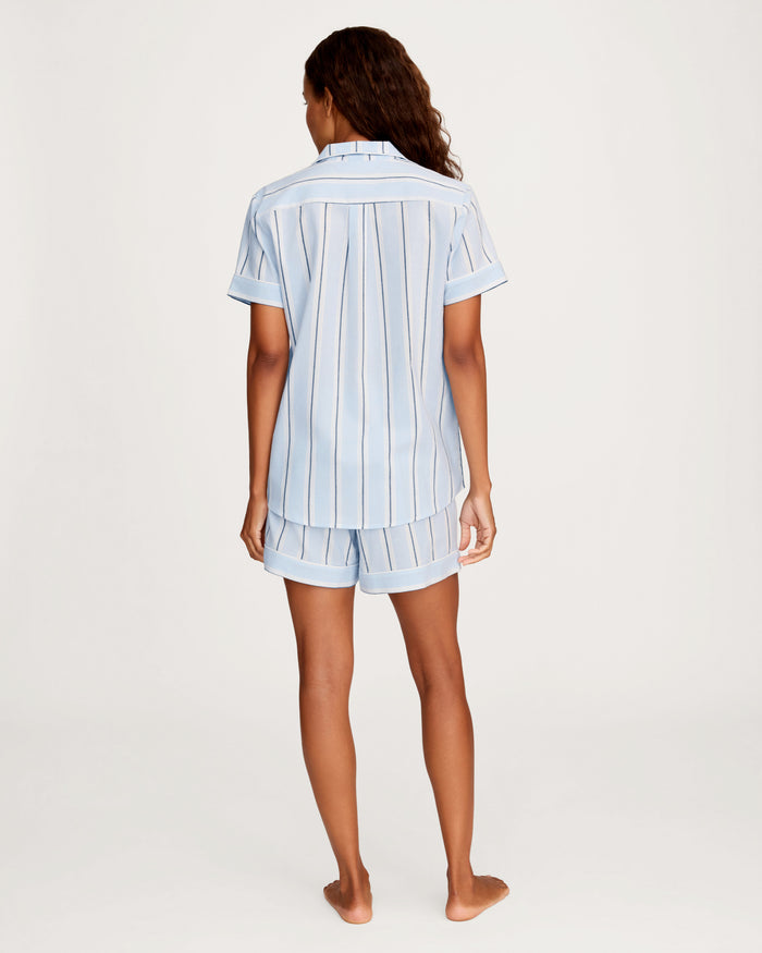 A woman stands barefoot, facing away, wearing the Women’s Twill Short Sleeve Short Set in Coastal Stripe—featuring light blue short-sleeved button-up pajamas with vertical white and dark stripes and matching shorts—against a plain light background.