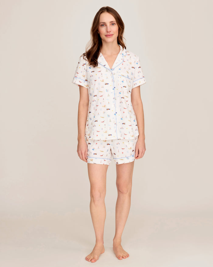 Against a plain light backdrop, a woman stands barefoot wearing the Women’s Twill Short Sleeve Short Set in Summer Nostalgia, featuring a matching short-sleeve pajama top and shorts adorned with a small, colorful print pattern on a light background.