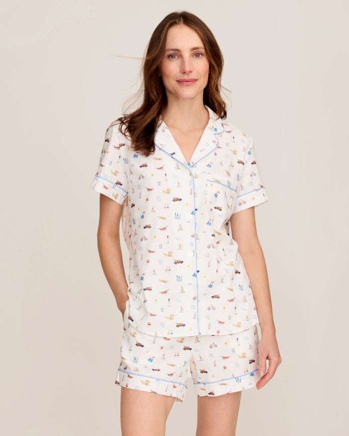A woman is wearing the Women’s Twill Short Sleeve Short Set in Summer Nostalgia, featuring a playful print with a short-sleeved, button-up top and matching shorts, as she stands smiling against a plain light background.