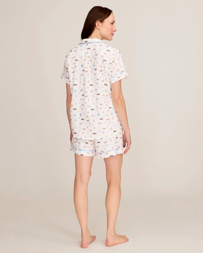 A woman stands barefoot, facing away, wearing the Women’s Twill Short Sleeve Short Set in Summer Nostalgia, featuring a white background with colorful prints. The scene is set against a simple, neutral backdrop.