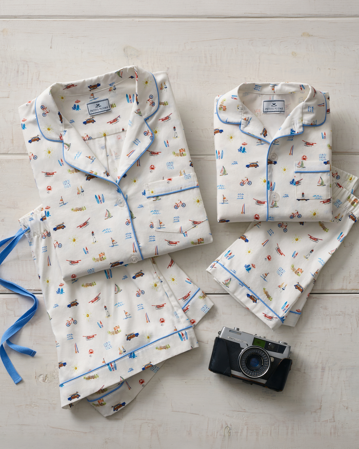 Two sets of the Kid’s Twill Pajama Short Set in Summer Nostalgia, featuring white fabric with colorful travel-inspired prints, are neatly folded on a white wooden surface. A vintage camera rests nearby, with a blue ribbon placed next to one pajama set.