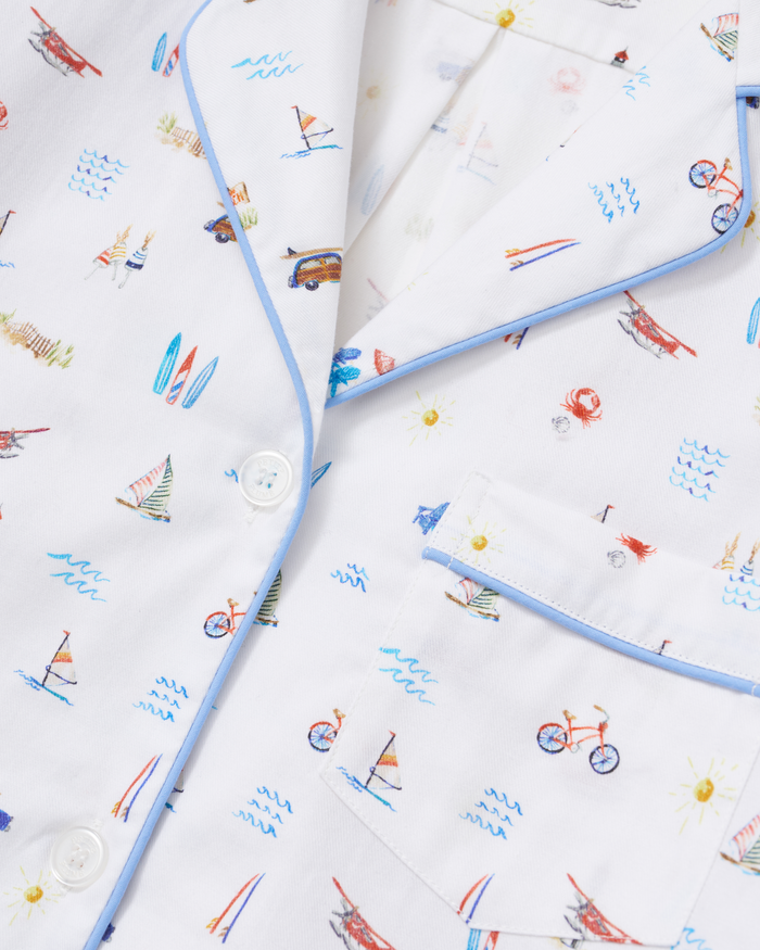 Women’s Twill Short Sleeve Short Set in Summer Nostalgia features a white shirt with a blue trim and white buttons, adorned with a playful summer pattern of colorful bicycles, sailboats, surfboards, cars, kites, suns, and blue waves. The shirt also includes a front pocket.