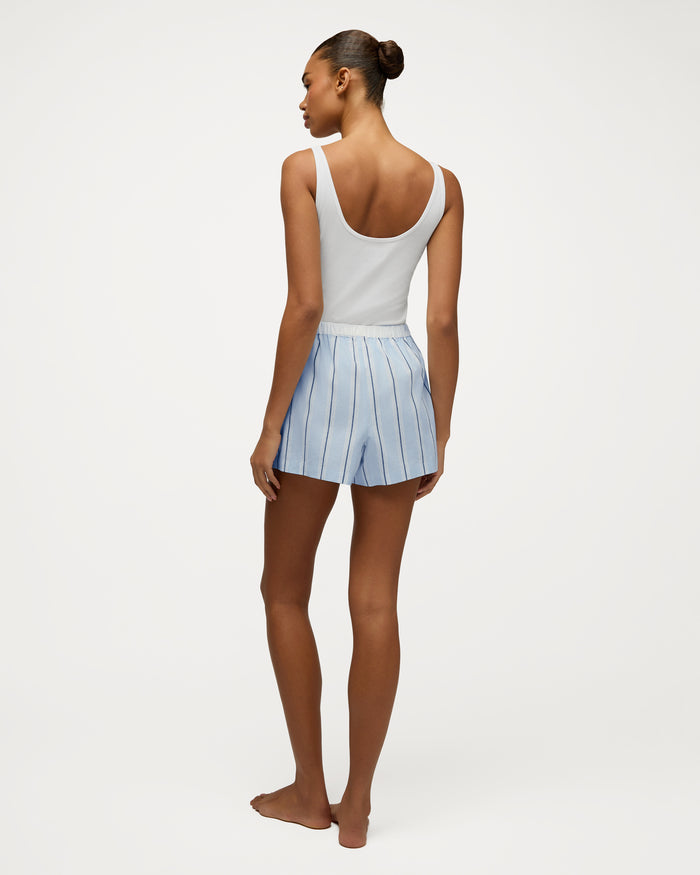 A woman stands barefoot facing away, wearing a white tank top and the Women’s Twill Sleep Short in Coastal Stripe. Her hair is tied back in a neat bun, and the background is plain white.