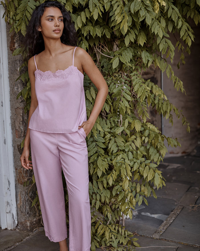 Wearing the Women’s Voile Fleur Cami Pant Set in Dusty Rose, a woman with long dark hair stands outdoors beside a leafy green vine, gazing to the side with one hand in her pocket.