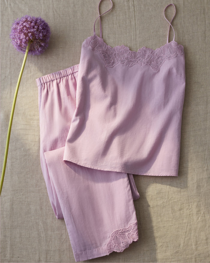 The Women’s Voile Fleur Cami Pant Set in Dusty Rose, featuring delicate lace detailing, is displayed on a beige fabric surface beside a single purple allium flower.