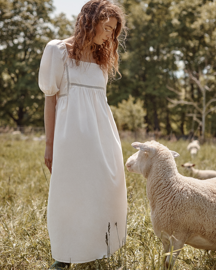 A woman wearing the Women’s Voile Lucienne Nightgown in Ivory stands in a grassy field, gazing down at a sheep. In the background, trees and more sheep can be seen on a sunny day.