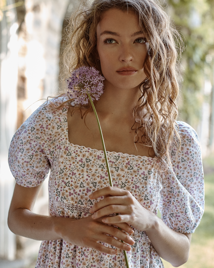 Wearing the Women’s Voile Lucienne Nightgown in Meadow Bloom, a woman stands outdoors, sunlight illuminating her wavy hair as she holds a purple allium flower and gazes thoughtfully into the distance.