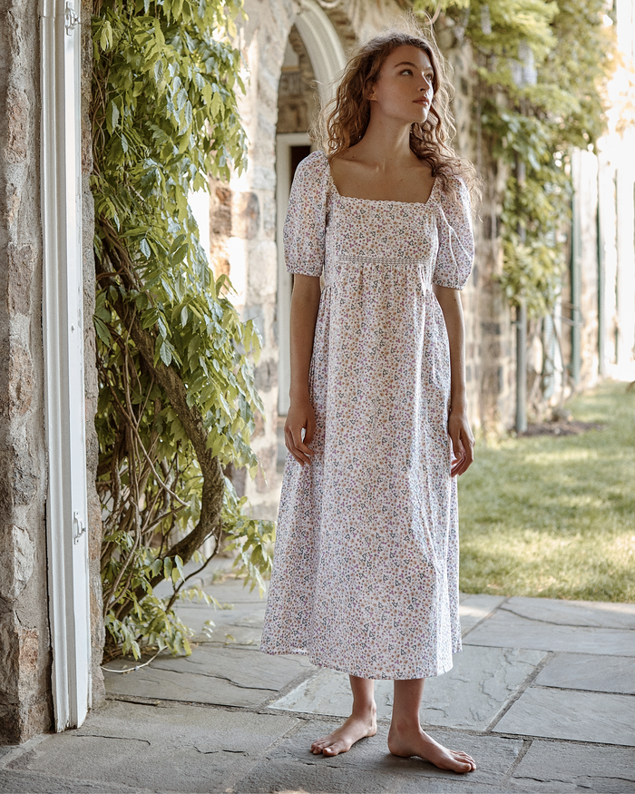 Wearing the Women’s Voile Lucienne Nightgown in Meadow Bloom, a barefoot woman stands on a stone path beside a vine-covered building, gazing thoughtfully into the distance.