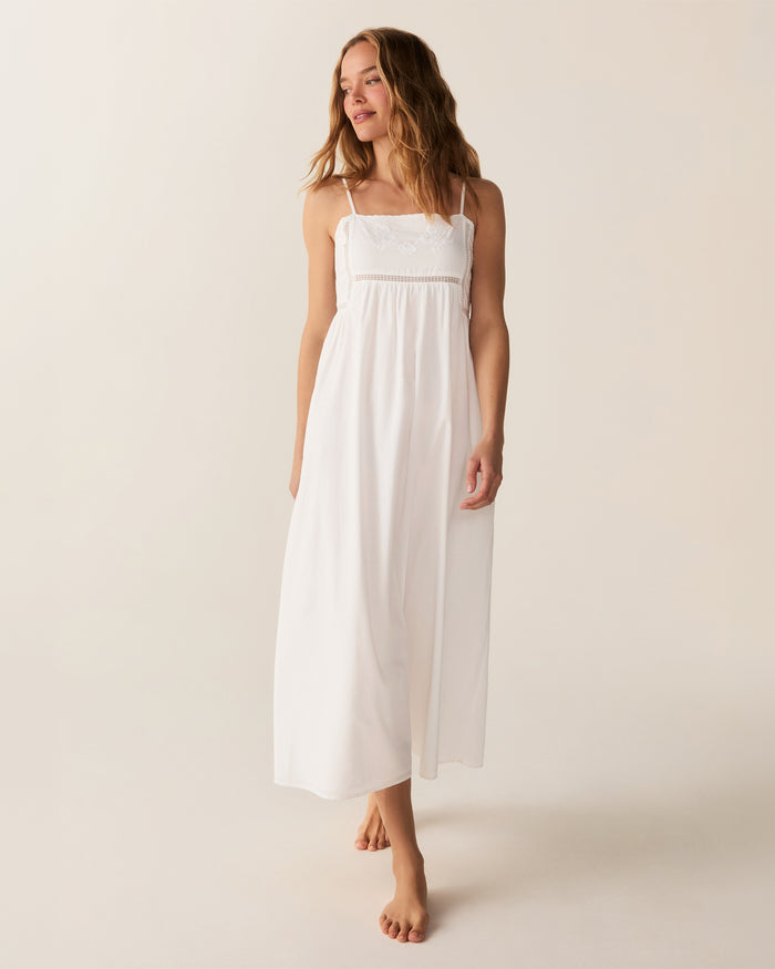 A woman with wavy hair stands barefoot and smiling to the side, wearing the Women's Voile Luna Gown in White—a long, sleeveless dress with thin straps—against a plain, light-colored background.