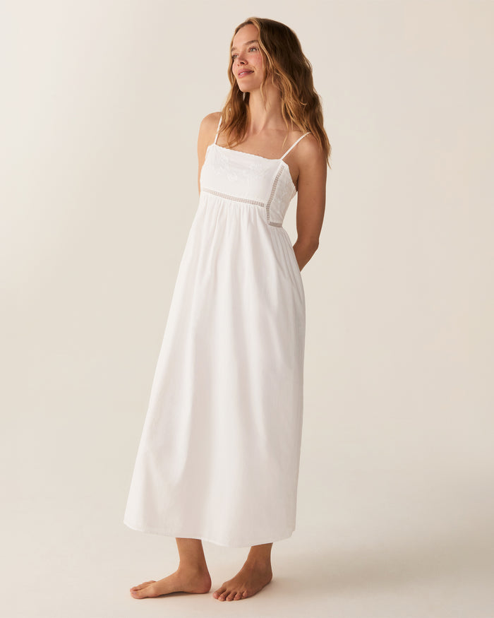 A woman with wavy hair stands barefoot and smiling to the side, wearing the Women's Voile Luna Gown in White—a long, sleeveless dress with thin straps—against a plain, light-colored background.