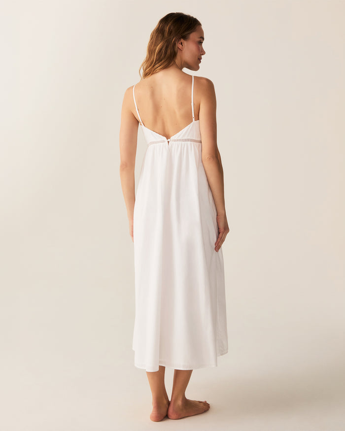 A woman with wavy hair stands barefoot and smiling to the side, wearing the Women's Voile Luna Gown in White—a long, sleeveless dress with thin straps—against a plain, light-colored background.