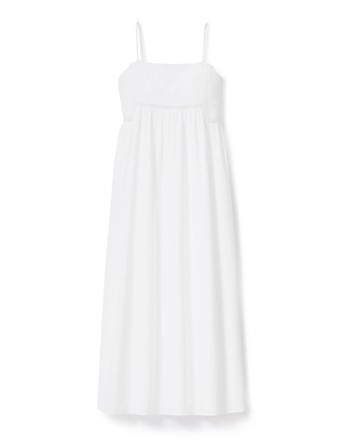 A woman with wavy hair stands barefoot and smiling to the side, wearing the Women's Voile Luna Gown in White—a long, sleeveless dress with thin straps—against a plain, light-colored background.