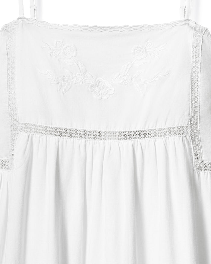 A woman with wavy hair stands barefoot and smiling to the side, wearing the Women's Voile Luna Gown in White—a long, sleeveless dress with thin straps—against a plain, light-colored background.